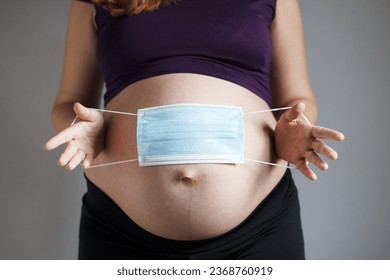 Close-up belly of pregnant woman and medical mask on a gray studio background, expectant mother and baby health protection concept, pregnancy and childbirth during infectious diseases - Shutterstock ID 2368760919
