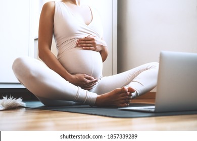 Closeup Belly Of Pregnant Woman Practicing Yoga Online At Home With Laptop. Expectant Mother Doing Prenatal Yoga Class Or Meditating During Pregnancy Indoors. Healthcare Of Mother And Unborn Baby.