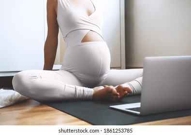 Closeup belly of pregnant woman practicing yoga online at home with laptop. Expectant mother doing prenatal yoga class or meditating during pregnancy indoors. Healthcare of mother and unborn baby.