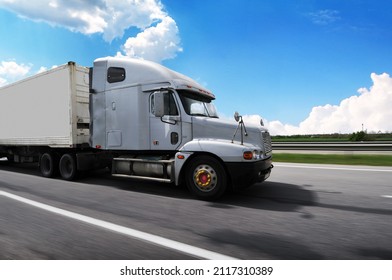 Close-up Of A Big Grey American Truck With A White Trailer Driving Fast On A Countryside Road Against A Blue Sky With Clouds