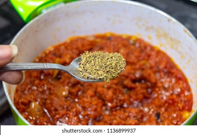 Closeup Of Black African American Cook's Hand Holding Spoon Of Spices And Thyme In The Tomato Sauce Pan On The Stove, Selective Focus, View From Above