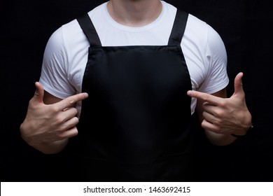 Closeup black blank empty apron on a strong guy chest. Fingers pointed to apron. Mock up, template for text design. Isolated on black background.