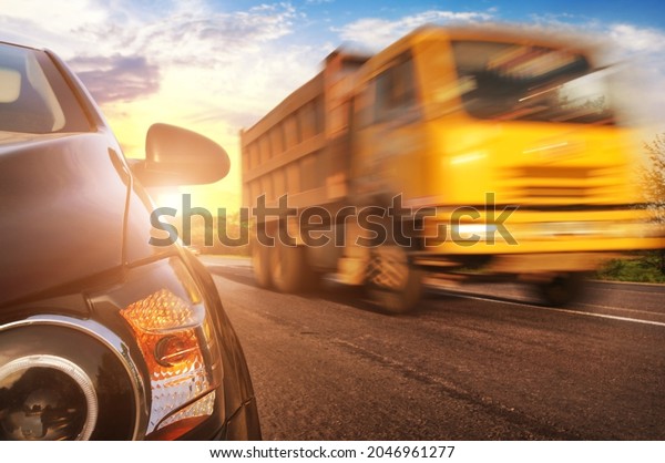 Close-up of a black car front end with\
a headlight and mirror on a countryside road with a yellow truck\
driving fast against a blue sky with a\
sunset