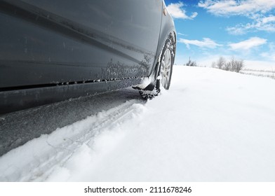 Close-up of a black car on a winter countryside road covered in snow against a blue sky with clouds