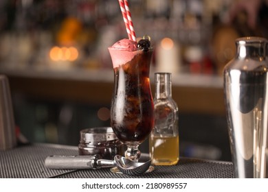 A Closeup Of Black Cherry Cocktail With Straw Near Glass Bottle And Spoon On Table