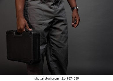 Close-up of a black construction suitcase in the strong male hands of a dark-skinned man in a gray jumpsuit or pants. A black guy with a watch on his arm carries a heavy suitcase with wrenches