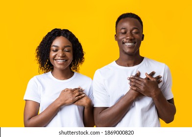 Closeup Of Black Couple Praying With Closed Eyes On Yellow Studio Background. Blessful African American Man And Woman Keeping Palms On Chest And Smiling, Cherishing Hope For Best