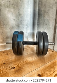 A close-up of a black dumbbell with blue padding resting on a wooden floor against a textured wall, creating a minimalistic fitness scene.