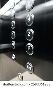 Close-up of black elevator control panel with illuminated metallic buttons showing floor numbers and parking level, modern lift interior design with reflective surface and chrome details.
