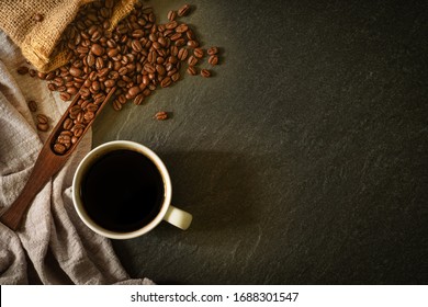 Close-up of Black hot coffee for morning with milk foam in white ceramic cup and napkin with coffee beans roasted in sack on dark table stone background. Top view, flat lay with copy space.