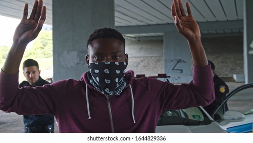 Close-up Black Man Perpetrator Wearing Mask Raising His Hands By Police Order. Police Officers On Duty Arrest An Afro-american Thief Bad Guy At Daytime.