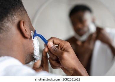 Closeup Of Black Man Shaving Face With Safety Razor Looking At Mirror Standing In Modern Bathroom At Home. Unrecognizable African American Guy Removing Hair On Face. Selective Focus - Powered by Shutterstock