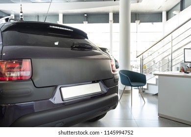 Close-up Of Black Mat Car In The Foreground And Office Desk With Chair In The Background Standing In Bright Car Showroom Interior With Windows