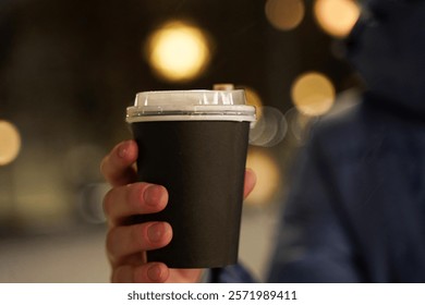 Close-up of black paper coffee cup in the hands of woman, standing on a cold street