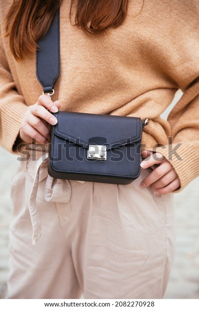 Closeup of black small handbag in woman\'s hand.\
Fall spring fashion outfit camel sweater and trendy white jeans.\
Fashion detail, stylish small bag. \
