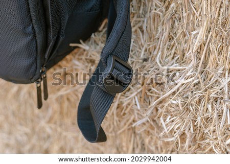 A close-up of the black strap of the backpack against the yellow hay. Black plastic buckle that adjusts the length of the backpack strap. Selective focus. Defocus, blur, noise, grain effect