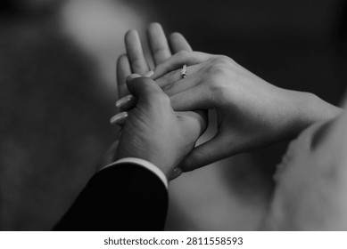 A close-up, black and white image capturing the intimate moment of a newly engaged or married couple holding hands, highlighting the sparkling diamond