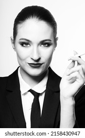 Close-up Black And White Portrait Of Woman Holding Cigarette In Her Hands Near Face And Looking To Camera. Model With Conservative Hairstyle And Wearing Black Classic Suit With Tie And White Shirt