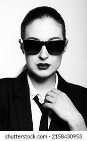 Close-up Black And White Studio Portrait Of Woman With Sunglasses And Suit Looking To Camera. Model With Conservative Hairstyle And Wearing Black Classic Suit With Tie And White Shirt