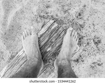 Closeup Black And White View Looking Down At A Mans Bare Feet And Toes Standing On A Piece Of Driftwood Plank Or Beam On A Sandy Beach.