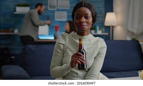 Closeup Of Black Woman Portrait Looking Into Camera Drinking Beer For Friend Birthday Celebration. In Background Group Of Mixed Race Friends Gathering Together Having Fun Late At Night In Living Room