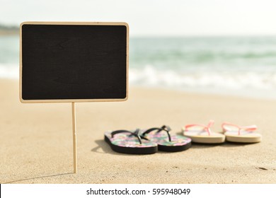 closeup of a blank black signboard and two pairs of flip-flops on the sand of a beach