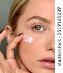 Close-up of a blonde woman with bright green eyes and freckles, applying moisturizer. She is wearing minimal makeup and wine red nail polish. This shot is capturing a daily self care +  beauty routine