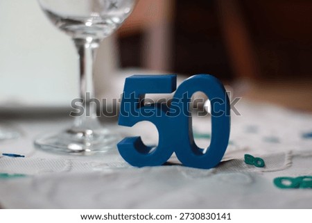 Close-up of a blue number 50 table decoration placed on an elegant dining table with a wine glass and light-colored tablecloth. The image represents a 50th birthday celebration, milestone anniversary, or festive event. Ideal for themes such as birthdays, anniversaries, family gatherings, hospitality, and elegant party decor.