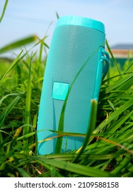 A Close-up Of A Blue Wireless Music Speaker Standing Among The Dense Green Grass. Modern Devices For Playing Music