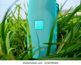 A Close-up Of A Blue Wireless Music Speaker Standing Among The Dense Green Grass. Modern Devices For Playing Music