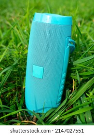 A Close-up Of A Blue Wireless Music Speaker Standing Among The Dense Green Grass. Modern Devices For Playing Music