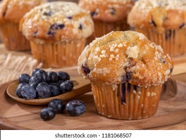 Closeup of a blueberry muffin and spoonful of berries on a wooden plate with muffins in background
