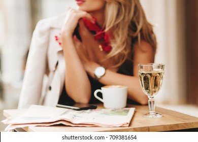 Close-up blur portrait of elegant woman wears red scarf and wristwatch with glass of wine on foreground. Blonde girl in stylish coat spending time in restaurant enjoying beverages and waiting friend.