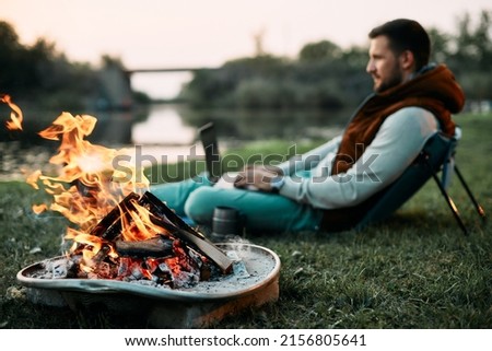 Close-up of bonfire with man working on laptop in the background in nature.