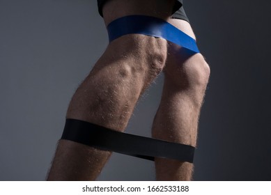 Closeup, Bottom View Of The Guys Muscular Legs In Sports Shorts, Stretching Fitness Elastic Bands With His Legs, Involvement Of Leg Muscle Groups That Are Not Involved In Everyday Life.