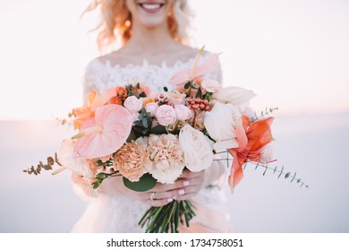 Close-up of a bride’s bouquet of amaryllis, anthurium, roses, carnations, eucalyptus in white-peach shades. The bride in a lace white dress holds a bouquet in her hands.  White sand