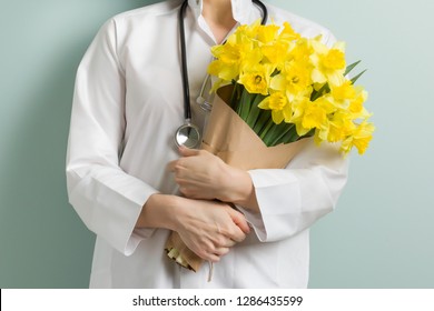 Closeup Of Bouquet Yellow Flowers In Hands Of Female Doctor, Pastel Green Mint Wall Background. World Health Day, Doctors Day.