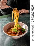 A close-up of a bowl of delicious noodle soup, with chopsticks lifting noodles above. Garnished with herbs and slices of meat, it