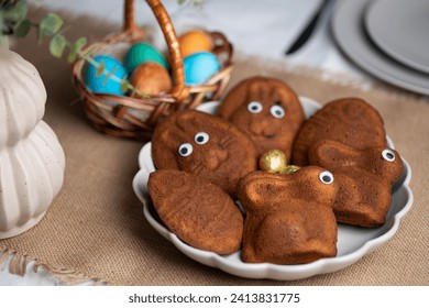 Close-up of bowl full of funny muffins shaped as bunnies and eggs with eyes. Multi colored Easter eggs in wicker basket and vase with green plant on served table