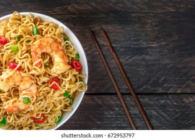 A closeup of a bowl of instant Chinese noodles with green onions, red hot chilli peppers, and shrimps, with chopsticks, shot from above on a dark rustic texture with a place for text - Shutterstock ID 672953797