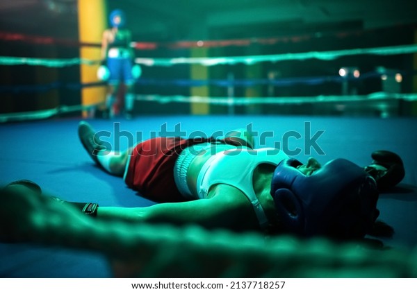 Close-up of boxer girl in knockout on ring. Two\
young girls boxing in gym when one girl fell down after took\
several blows of partner and lying on floor. Active lifestyle,\
hobby, womens boxing\
concept