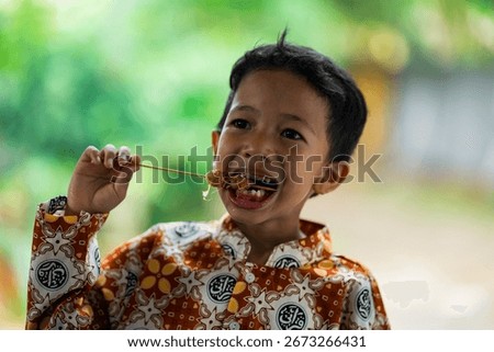 Close-up of a boy smiling while eating satay on a stick, enjoying delicious food outdoors with happiness.	
