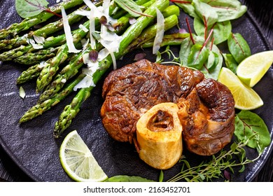 close-up of braised osso buco, veal shank steak  with grilled asparagus, lime and swiss chard fresh leaves on black plate on dark wood table