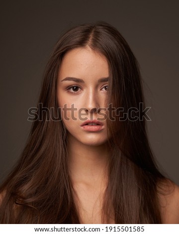 Close-up breast-up portrait of young beautiful brunette teenager girl with long straight hair and perfect natural make-up posing against grey background