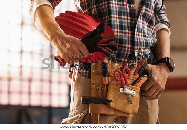 Closeup of bricklayer hands holding hardhat\
and construction equipment. Detail of mason man hands holding work\
gloves and wearing tool kit on waist. Handyman with tools belt and\
artisan equipment.
