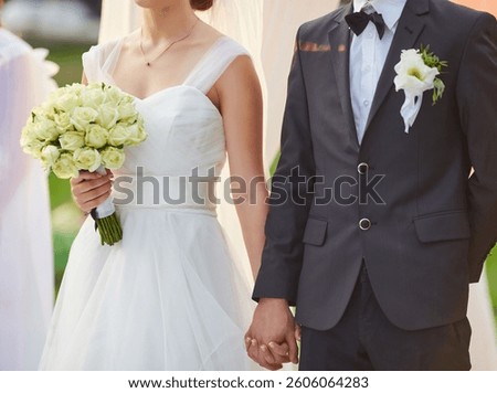 Close-up of a bride holding a white rose bouquet and the groom s hand during their wedding ceremony, highlighting the elegant wedding dress and groom s suit with a boutonniere.