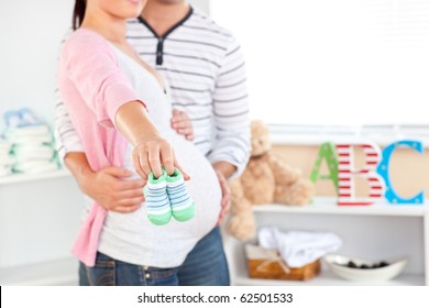 Close-up of a bright pregnant woman holding baby shoes while husband touching her belly in the room of their future baby at home