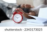 Close-up of bright red alarm clock and young latino woman turning it off while laying in bed. Ten minutes to eleven. Book pages on table near wake-up call