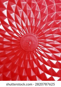 A close-up of a bright red plastic surface with a radial geometric pattern. The design features repeated triangular facets that radiate outward from a small circular center, creating a sunburst or star-like effect. The glossy material reflects light, giving highlights and shadows that emphasize the texture and depth