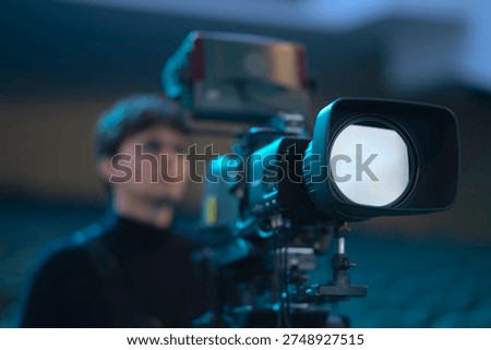 Close-up of a broadcasting camera in a blue studio hall. Young man silhouette on the background. Concept of media production, future of television, and cinematography